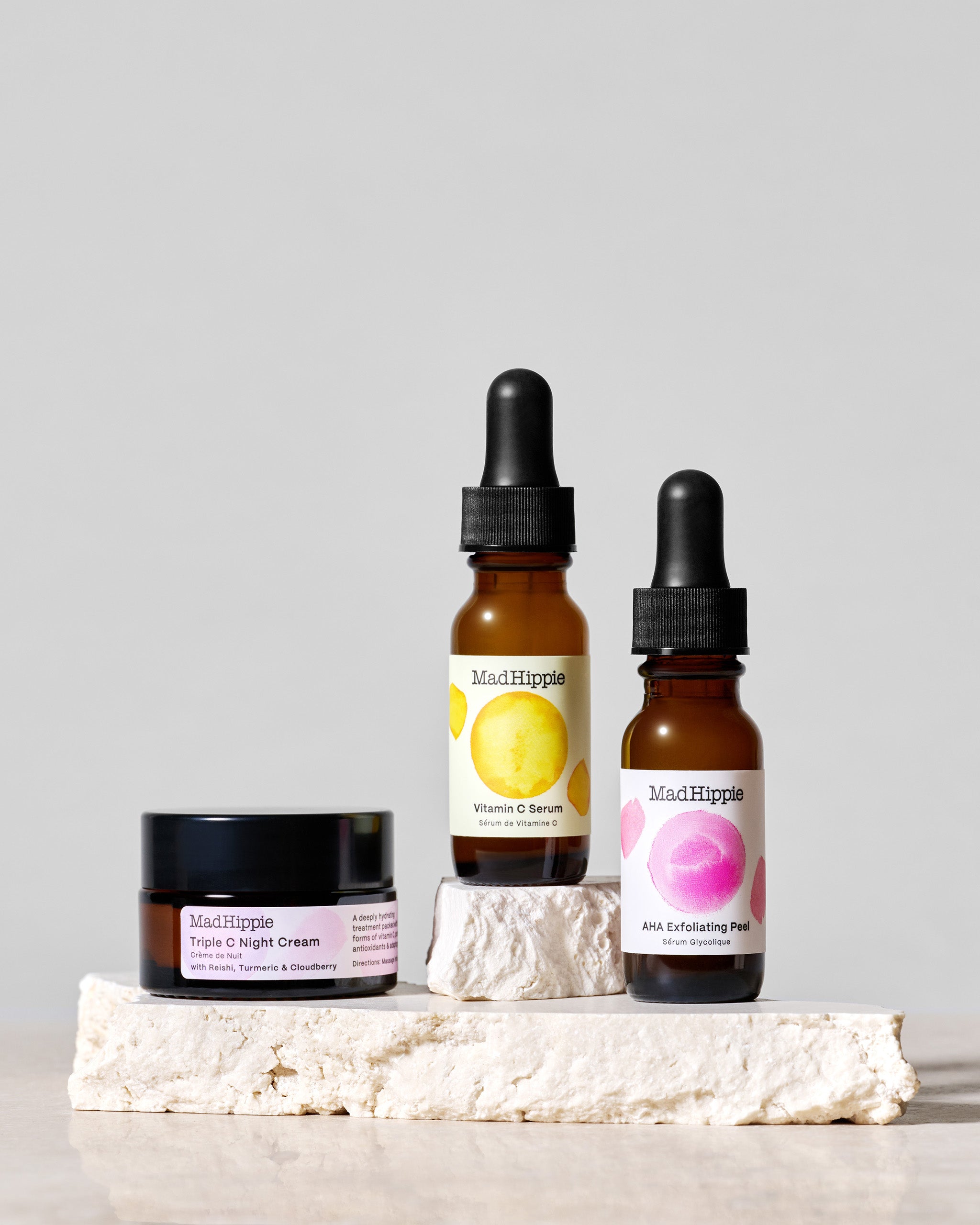 Triple C Night Cream jar + Vitamin C Serum and AHA Exfoliating Peel bottles on stone slabs, with gray background